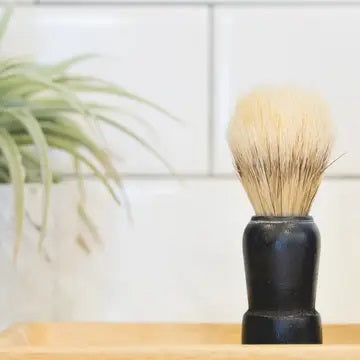 THE IMMACULATE BEARD - SHAVE BRUSH by THE IMMACULATE BEARD, featuring a black handle and faux boar bristles, stands upright on a wooden tray with a green plant and white tiled wall blurred in the background.