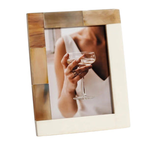 Two luxury ZODAX photo frames—one crafted from natural white horn inlay and the other an elegant ivory resin—display portraits: one shows a person holding a glass, while the other features a close-up of a face with a finger touching the lips. Both WHITE HORN AND IVORY RESIN FRAME - 5X7 rest on a patterned surface.