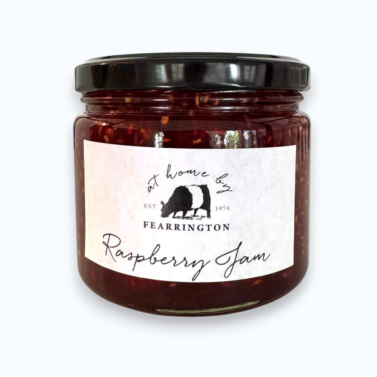 A glass jar of raspberry jam is displayed with a black and white striped ribbon adorning the lid. The white label reads "Fearrington Housemade Raspberry Jam" with a list of ingredients: raspberry, vanilla, lemon, pectin, sugar. The text also indicates it is handmade and may contain allergens. Perfect as a hostess gift!