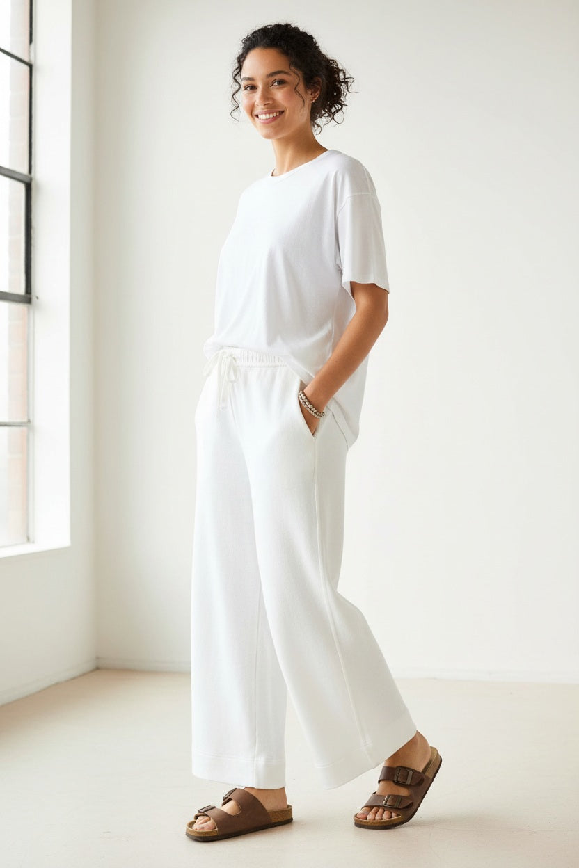 A person stands on a sidewalk in front of urban buildings and a street, wearing a white sweatshirt paired with the MERSEA - FRENCHIE WIDE LEG PANT IN PERIWINKLE from MERSEA. The relaxed fit French terry pants feature a wide-leg silhouette and are styled with sporty beige-and-blue sneakers and a cream tote bag with dark trim.