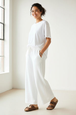 A person stands on a sidewalk in front of urban buildings and a street, wearing a white sweatshirt paired with the MERSEA - FRENCHIE WIDE LEG PANT IN PERIWINKLE from MERSEA. The relaxed fit French terry pants feature a wide-leg silhouette and are styled with sporty beige-and-blue sneakers and a cream tote bag with dark trim.