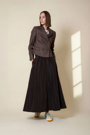 A person wearing a white sweater has their hand in the pocket of the VICARIO CINQUE GRANADA Taffeta Skirt in Marron Brown, a high-waisted skirt crafted from memory taffeta with a subtle sheen.