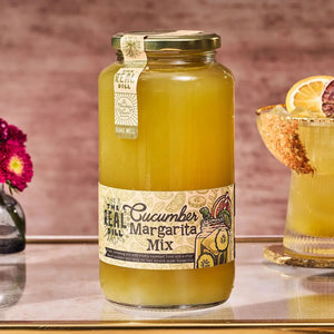 A glass jar of THE REAL DILL - CUCUMBER MARGARITA BLOODY MARY MIX 32OZ features a light green-yellow liquid and a decorative label with cucumber and citrus slice illustrations; this fresh-squeezed cocktail mixer from THE REAL DILL provides a low sugar margarita experience perfect for any occasion.