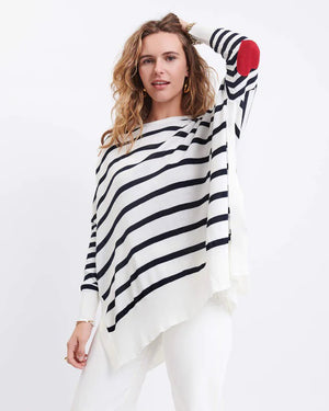 A woman with wavy hair smiles in the MERSEA - AMOUR HEART SWEATER, a French-inspired striped sweater with a red heart elbow patch, paired with white pants against a plain white background.