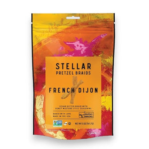 A vibrant bag in shades of orange and yellow—labeled “STELLAR SNACKS FRENCH DIJON PRETZEL BRAIDS”—features a vegan butter braid with honey mustard style seasoning. The packaging highlights that the product is Non-GMO, and “Baked with Love” is prominently displayed at the bottom.