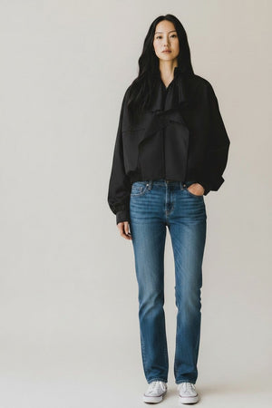A woman faces forward against a plain background, wearing a black top, blue jeans, white sneakers, and the NUDE Lightweight Bomber Jacket with Ruffle by NUDE draped over her shoulders. Her dark hair is loose and she looks relaxed.