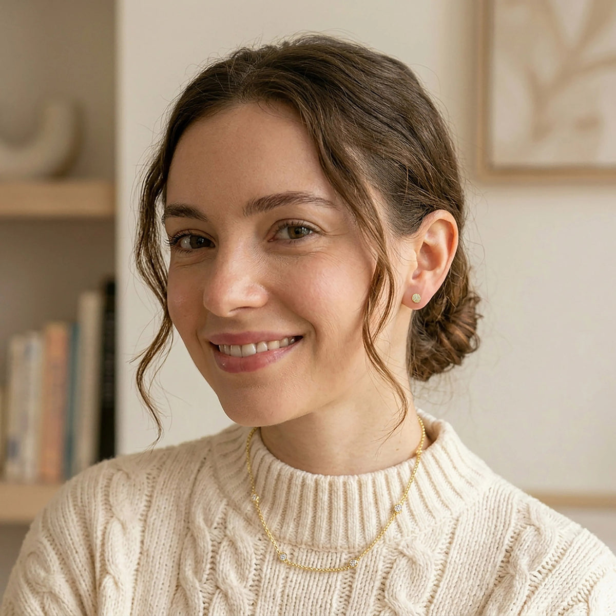 A woman with brown hair in a low bun, wearing a cream cable-knit sweater and ELLA STEIN - ROUND STUD EARRINGS by ELLA STEIN, smiles softly. She stands indoors with shelves and a framed picture blurred in the background.