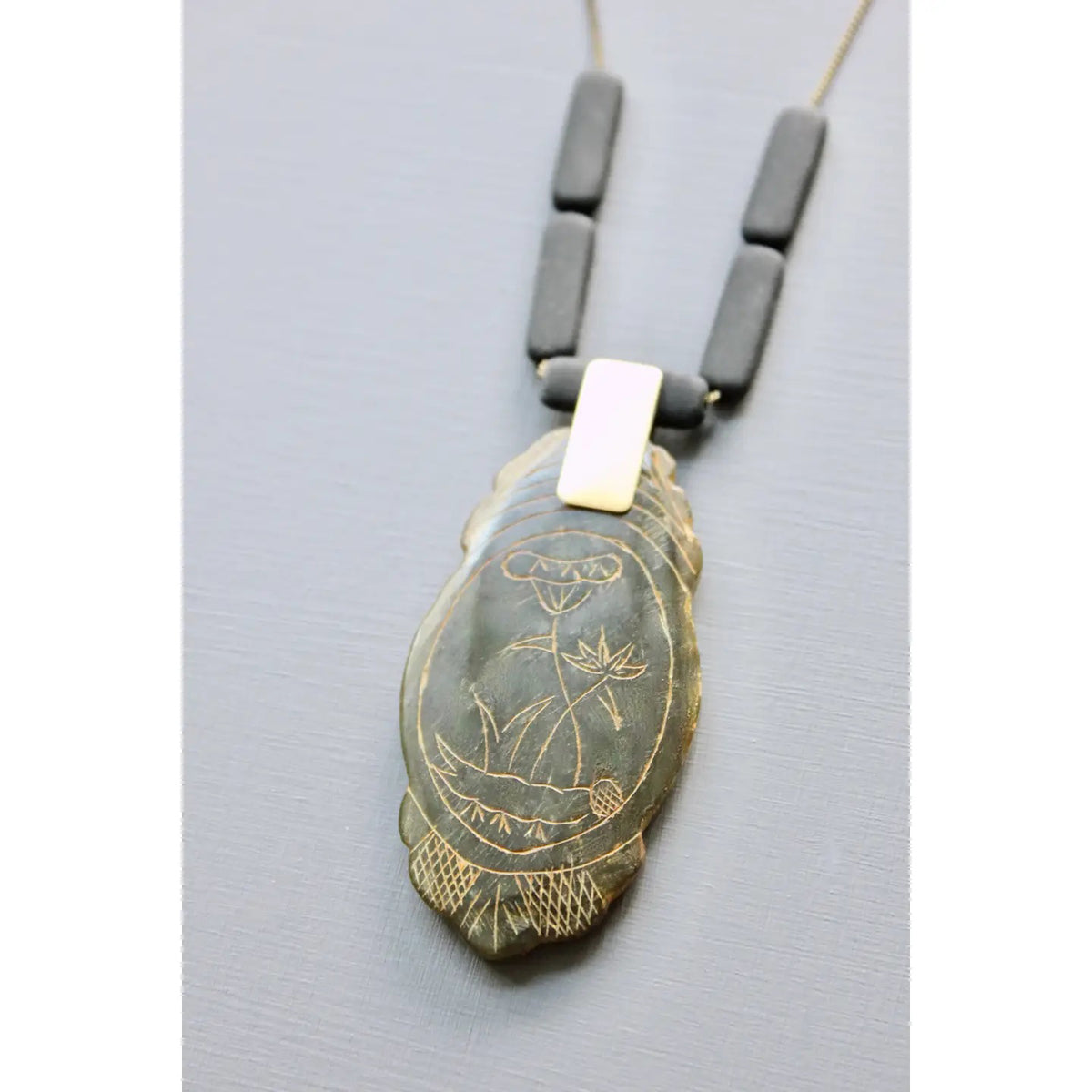 The DAVID AUBREY - CARVED JADE PENDANT NECKLACE by DAVID AUBREY features a 24-inch brass-ox rectangular chain with three black beads and a feather-shaped pendant engraved with an owl and plant design, displayed on a light grey background.
