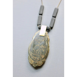 The DAVID AUBREY - CARVED JADE PENDANT NECKLACE by DAVID AUBREY features a 24-inch brass-ox rectangular chain with three black beads and a feather-shaped pendant engraved with an owl and plant design, displayed on a light grey background.