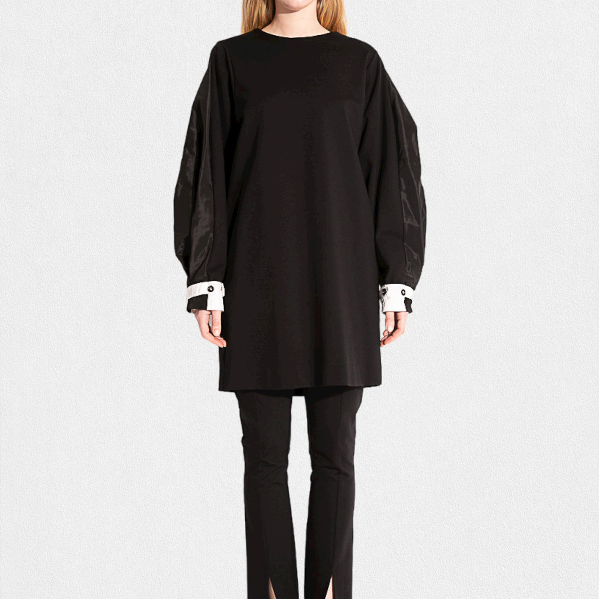 Dressed in the MEIMEIJ Knit and Taffeta Tunic/Dress, a versatile black piece with flowing sleeves and white cuffs, the ensemble is elegantly completed with black pants featuring subtle ankle slits, standing out against a plain white backdrop.