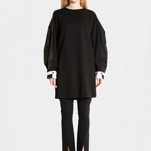 Dressed in the MEIMEIJ Knit and Taffeta Tunic/Dress, a versatile black piece with flowing sleeves and white cuffs, the ensemble is elegantly completed with black pants featuring subtle ankle slits, standing out against a plain white backdrop.