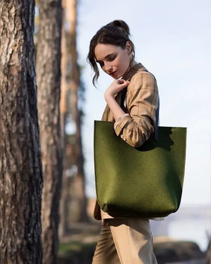 A person in a beige trench coat holds the WOOLBERRY WOOL FELT LONG TOTE BAG with black handles, standing against a plain, light-colored background.
