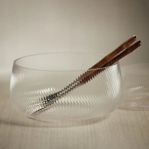 The CONNAUGHT RIPPLED GLASS BOWL - LARGE by ZODAX is an exquisite piece with a clear, textured design featuring a subtle diagonal stripe pattern. Measuring 10.75 inches in diameter and 5 inches in height, this bowl boasts a rounded shape with a wide, open top. Placed on a reflective white surface, its elegant design and transparency are beautifully highlighted, emphasizing its sophisticated dimensions.