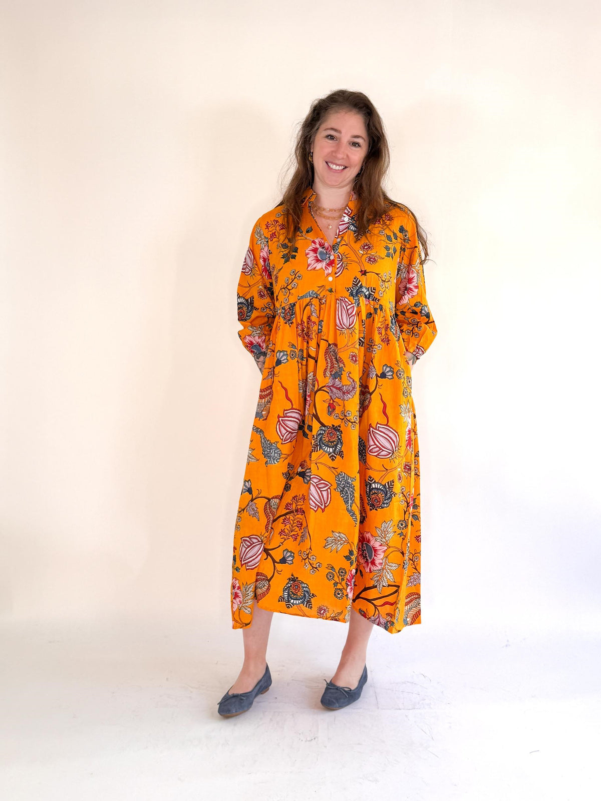 A woman smiles against a plain background, wearing FARYLINA&#39;s MAPLE LONG DRESS—a bright orange, flowy cotton midi with a bold floral pattern—and blue flats, her hands behind her back.