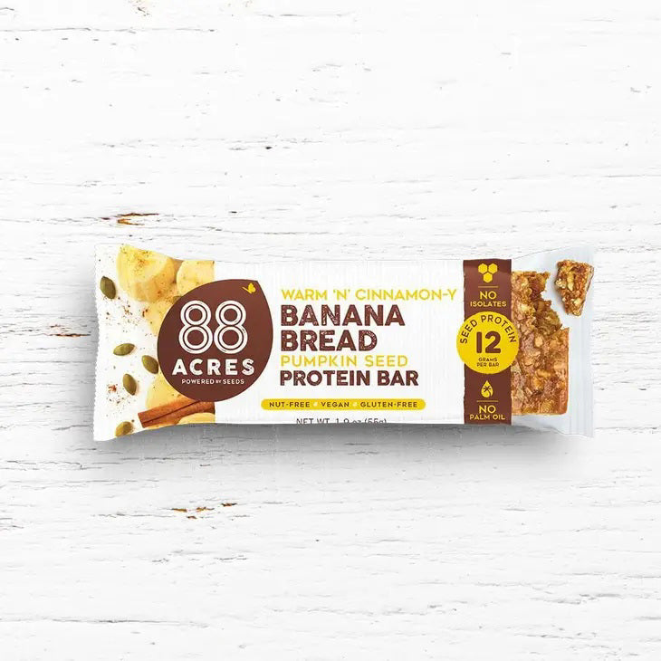 A protein bar wrapper labeled &quot;88 ACRES - BANANA BREAD HIGH PROTEIN BAR&quot; by 88 Acres. The wrapper highlights features such as &quot;Nut-Free,&quot; &quot;Vegan,&quot; &quot;Gluten-Free,&quot; 12 grams of protein, and &quot;No Palm Oil.&quot; The wrapper is set against a white wooden background.