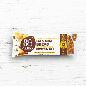 A protein bar wrapper labeled "88 ACRES - BANANA BREAD HIGH PROTEIN BAR" by 88 Acres. The wrapper highlights features such as "Nut-Free," "Vegan," "Gluten-Free," 12 grams of protein, and "No Palm Oil." The wrapper is set against a white wooden background.