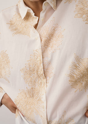 A woman with straight blonde hair and a neutral expression stands against a plain light wall, wearing the 0039 ITALY LUCY BLOUSE WITH EMBROIDERED FLOWERS, light blue wide-leg jeans, and beige flat shoes.