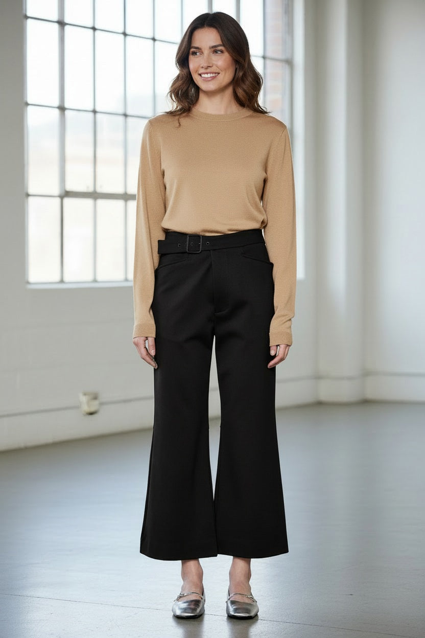 A person models the MEIMEIJ Crop Ponte Pant with Belt, shown in brown with pockets, paired with a beige top and silver flats. Only the lower half is visible against a plain white wall and concrete floor.