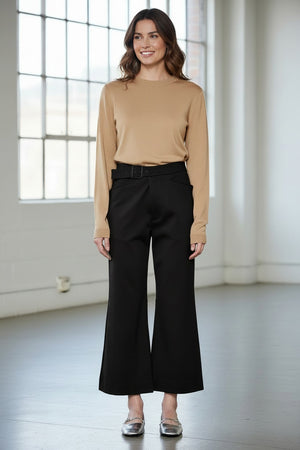 A person models the MEIMEIJ Crop Ponte Pant with Belt, shown in brown with pockets, paired with a beige top and silver flats. Only the lower half is visible against a plain white wall and concrete floor.