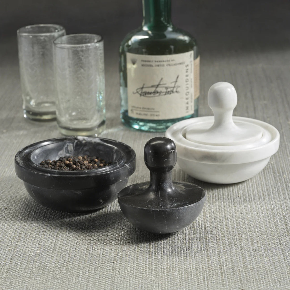 ZODAX MARBLE SPICE GRINDER in black and white rests on a gray surface, one containing black peppercorns. Two textured glasses and a green bottle in the background emphasize the elegant marble design of this kitchen essential.
