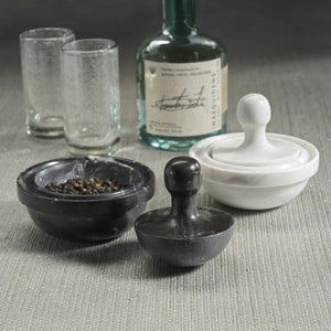 ZODAX MARBLE SPICE GRINDER in black and white rests on a gray surface, one containing black peppercorns. Two textured glasses and a green bottle in the background emphasize the elegant marble design of this kitchen essential.