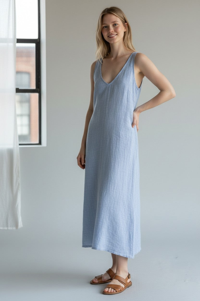 A woman stands indoors wearing the LLEA BLUE - ERICA Linen Bias Cut Dress, a sleeveless, light blue design from LLEA BLUE, paired with brown sandals. She smiles with one hand on her hip, while a window and sheer curtain can be seen in the background.
