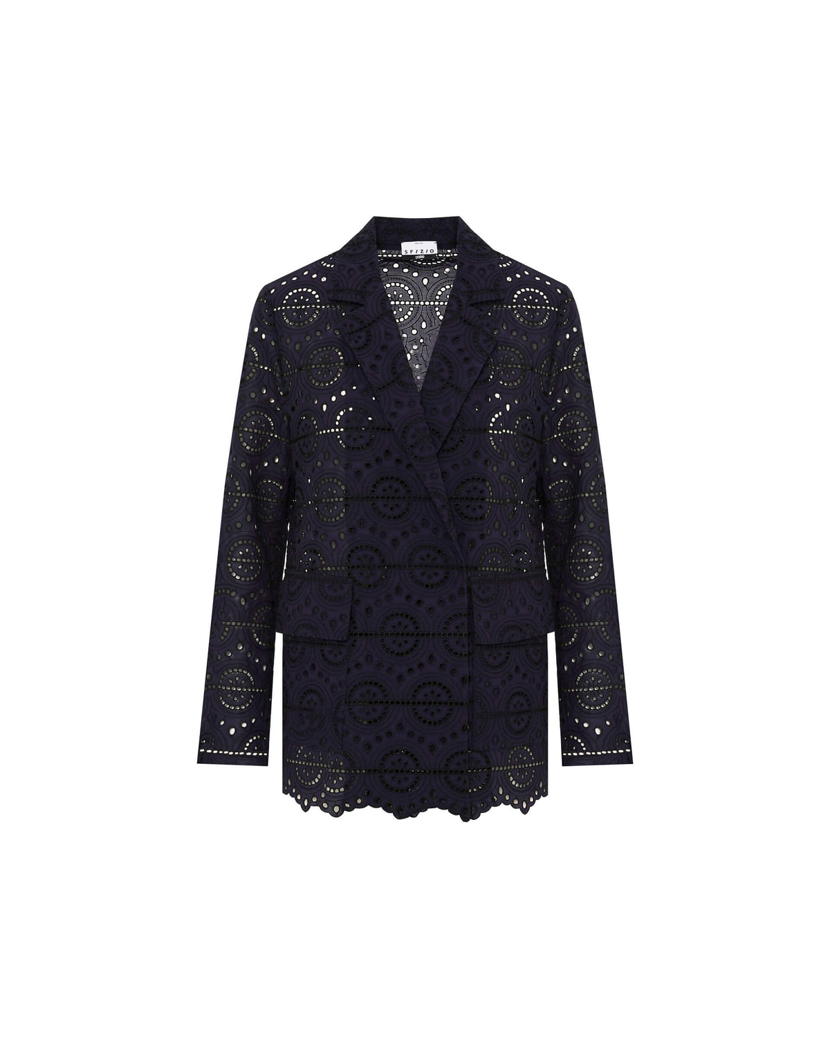 A person with long, straight hair stands against a pale yellow background, donning the SFIZIO Double Breasted Broderi Anglaise Jacket by SFIZIO. The intricate circular patterns on the dark navy lace pantsuit add an elegant touch. The double-breasted jacket is buttoned up, and the person wears a serious expression with minimal accessories.