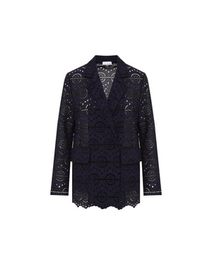 A person with long, straight hair stands against a pale yellow background, donning the SFIZIO Double Breasted Broderi Anglaise Jacket by SFIZIO. The intricate circular patterns on the dark navy lace pantsuit add an elegant touch. The double-breasted jacket is buttoned up, and the person wears a serious expression with minimal accessories.