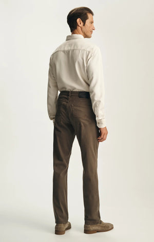 A person is seen from behind, standing against a plain light background, wearing the 34 HERITAGE - CHARISMA RELAXED STRAIGHT PANTS IN HARDWOOD TWILL by 34 HERITAGE. The brown comfort stretch twill pants are paired with a cream long-sleeve shirt and brown shoes.