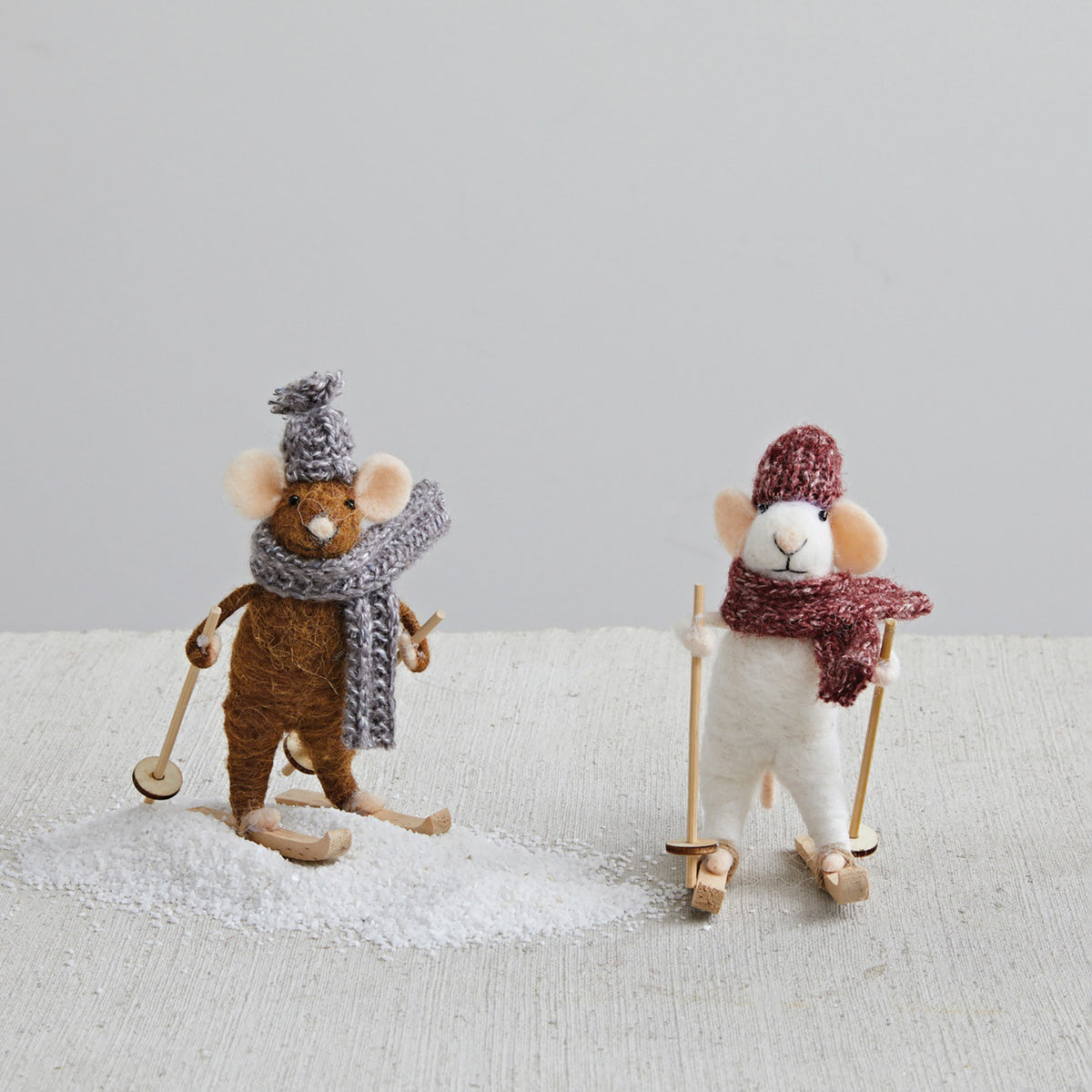 The CREATIVE COOP FELT SKIING MOUSE features one brown and one white mouse, each in knitted hats and scarves, standing on skis with poles. They showcase assorted cozy accessories atop a textured white surface.