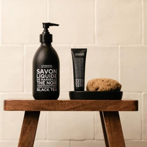A black tube of COMPAGNIE DE PROVENCE - TRAVEL HAND CREAM BLACK TEA 1OZ from CIE LUXE BRANDS stands upright on a light, cylindrical stone pedestal against a neutral beige background with soft lighting.