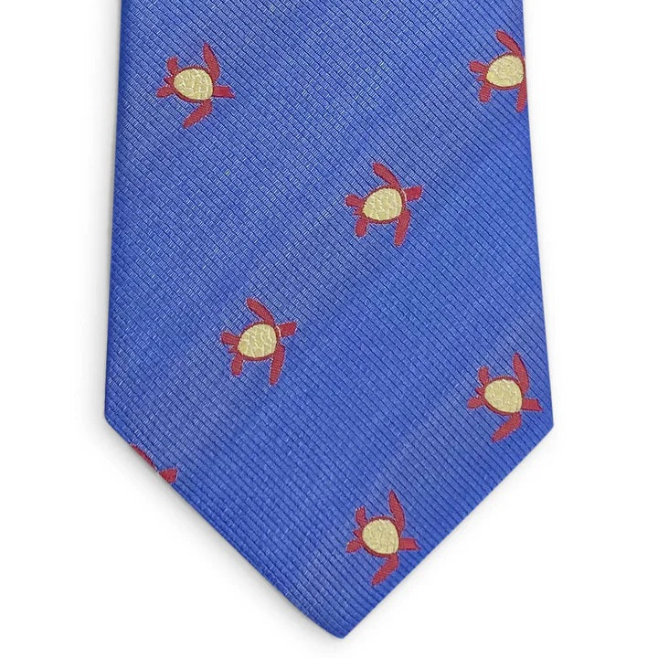 A rolled-up blue silk tie from BIRD DOG BAY, named the PETER-BLAIR - TURTLES SILK TIE IN BLUE, features a repeating yellow and red sea turtle pattern.