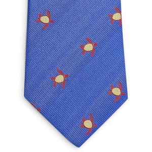 A rolled-up blue silk tie from BIRD DOG BAY, named the PETER-BLAIR - TURTLES SILK TIE IN BLUE, features a repeating yellow and red sea turtle pattern.