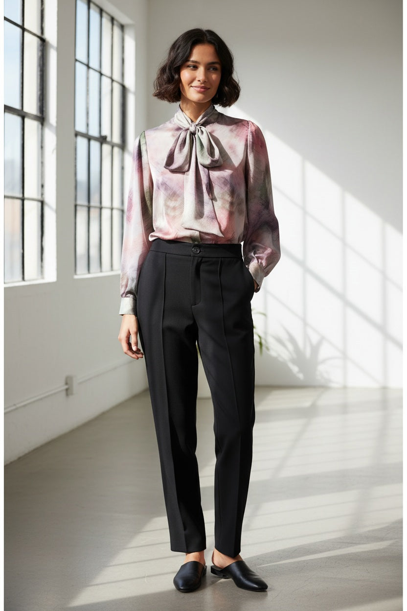 A woman with short dark hair wears the MILVA MI - BRUSHSTROKE BLOUSE by MILVA, a pastel long-sleeved statement bow top, paired with high-waisted black pants. She poses with eyes closed against a plain light background.