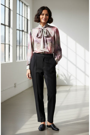 A woman with short dark hair wears the MILVA MI - BRUSHSTROKE BLOUSE by MILVA, a pastel long-sleeved statement bow top, paired with high-waisted black pants. She poses with eyes closed against a plain light background.