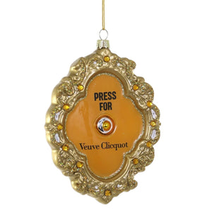 A 3.5-inch oval-shaped Cody Foster PRESS FOR FRENCH CHAMPAGNE ORNAMENT in a gold frame, featuring a center button, small inlaid jewels, and text that reads “PRESS FOR Veuve Clicquot.”.