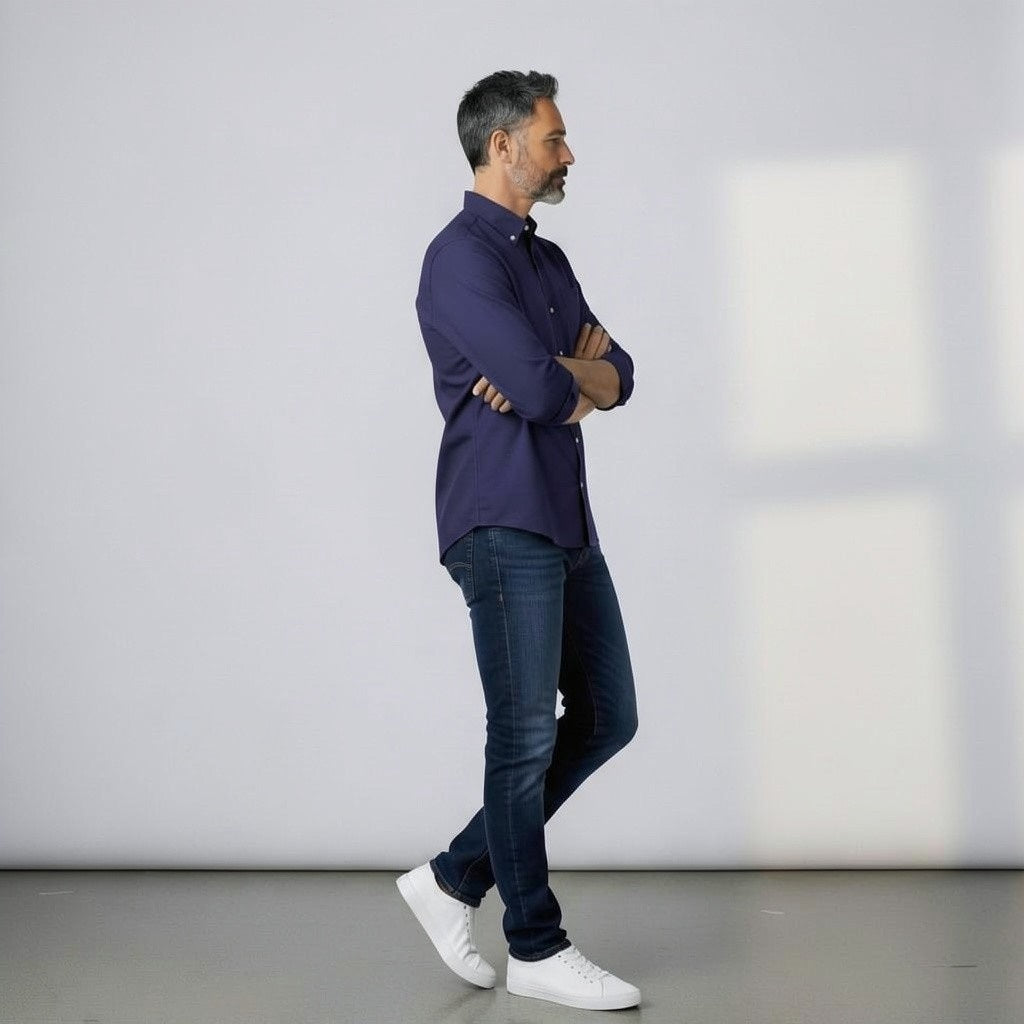 A man with short dark hair and a beard stands sideways with arms crossed, wearing the FRANK & EILEEN LUKE Men's Button Up Shirt in Famous Denim Washed Navy, blue jeans, and white sneakers. He is indoors by a light wall.