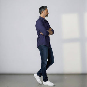 A man with short dark hair and a beard stands sideways with arms crossed, wearing the FRANK & EILEEN LUKE Men's Button Up Shirt in Famous Denim Washed Navy, blue jeans, and white sneakers. He is indoors by a light wall.