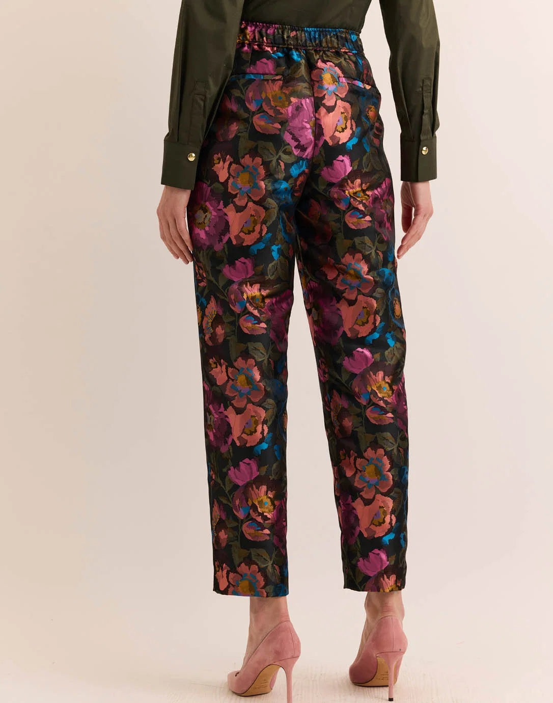 A person wears the HINSON WU Jolene Jacquard Pant in Floral Bouquet—high-waisted, ankle-length tailored trousers featuring a vibrant floral pattern in pink, purple, blue, and orange—paired with a dark green shirt and light pink heels.