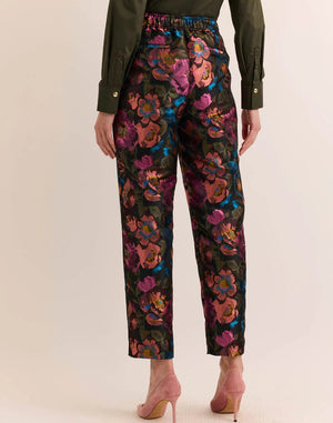 A person wears the HINSON WU Jolene Jacquard Pant in Floral Bouquet—high-waisted, ankle-length tailored trousers featuring a vibrant floral pattern in pink, purple, blue, and orange—paired with a dark green shirt and light pink heels.