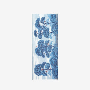 A woman in a sleeveless beige dress faces forward, draped in the INOUI EDITIONS - AMBROISE SCARF IN DUCK BLUE by INOUI, featuring a dark blue botanical pattern on linen cotton. The background is plain white.
