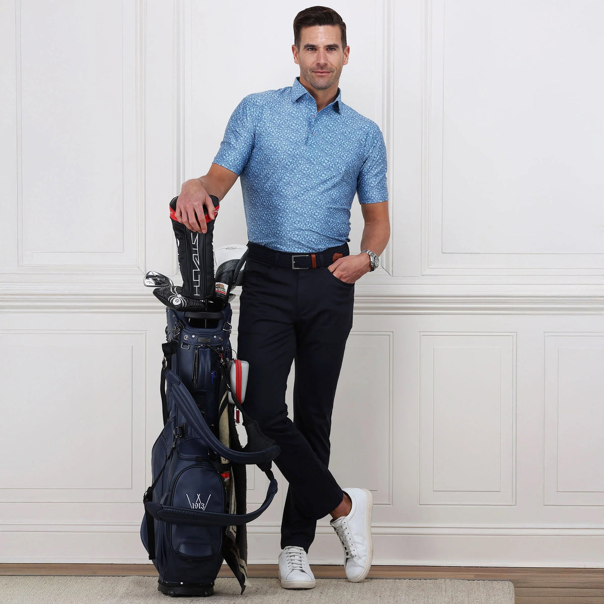 A man wearing a blue patterned polo, COLLARS AND CO Velocity Pull-On Performance Pants in Navy, and white sneakers stands indoors, confidently leaning on a golf bag against a white paneled wall.
