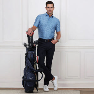 A man wearing a blue patterned polo, COLLARS AND CO Velocity Pull-On Performance Pants in Navy, and white sneakers stands indoors, confidently leaning on a golf bag against a white paneled wall.
