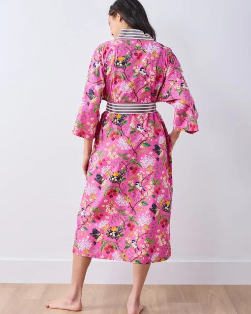 A person wearing a PRINTFRESH - BIRD & BLOSSOM LONG ROBE IN FUSCHIA, a long organic cotton robe with a bird and blossom pattern and green and white striped trim.