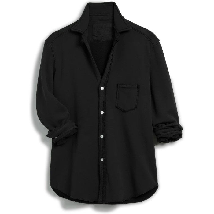 A person with curly hair smiles while standing next to a wooden table, wearing the EILEEN Relaxed Sweatshirt Button Up by FRANK &amp; EILEEN, along with white pants. In the background are wooden panels, framed botanical artwork, and a piano adorned with flowers, creating a serene scene.