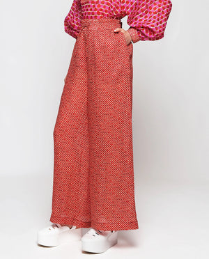 A person wearing the MIRTO 1956 - MOSAIC RAMIE TROUSER, featuring a high-waisted, wide-leg design with an elastic waistband and a small geometric pattern. The trousers are paired with a matching patterned top with billowy sleeves from MIRTO 1956. Completing the stylish yet comfortable look, the person is also wearing white platform sneakers.