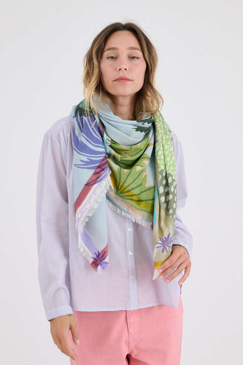 A woman smiles while holding the STORIATIPIC - CALI MAX SCARF, showcasing its vibrant tropical print of palm trees and boats. She pairs this luxury accessory from STORIATIPIC with a light blouse, pink pants, and green shoes.