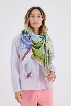 A woman smiles while holding the STORIATIPIC - CALI MAX SCARF, showcasing its vibrant tropical print of palm trees and boats. She pairs this luxury accessory from STORIATIPIC with a light blouse, pink pants, and green shoes.
