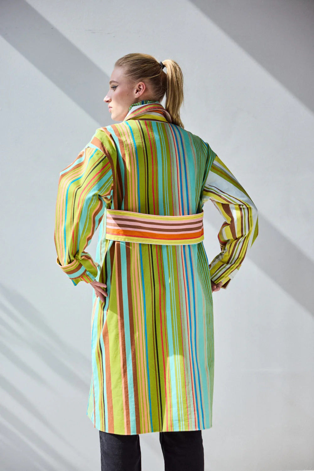 A person stands against a white wall wearing the HOPE FOR FLOWERS - COMBO STRIPES COAT by HOPE FOR FLOWERS, styled over a patterned shirt and black pants. Sunlight casts leaf-like shadows on the wall and floor behind them.