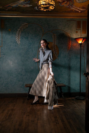 A woman stands in a dimly lit, ornately decorated room, wearing the HOPE FOR FLOWERS - CIRCLE SKIRT IN NATURAL CHECK by HOPE FOR FLOWERS. Warm light from a floor lamp casts shadows on the teal wall behind her.